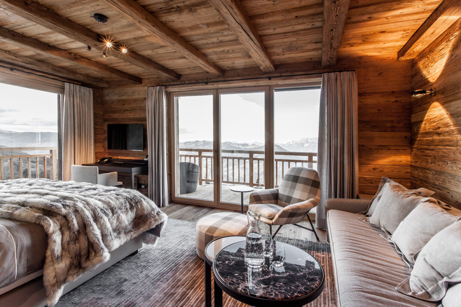 Cozy modern bedroom with wooden beams, featuring a plush bed with a fur throw, a comfortable sofa, a sleek coffee table, and large windows offering scenic mountain views. | Only on SopranoVillas