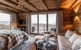 Cozy modern bedroom with wooden beams, featuring a plush bed with a fur throw, a comfortable sofa, a sleek coffee table, and large windows offering scenic mountain views.