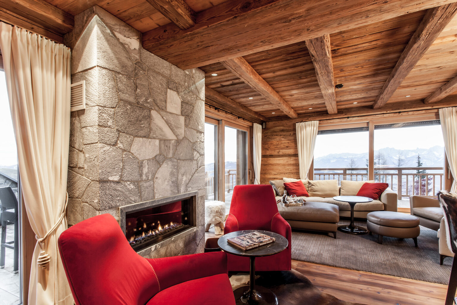 Cozy living room with red chairs, a stone fireplace, and large windows offering mountain views, featuring wooden beams and soft curtains for a warm ambiance. | Only on SopranoVillas