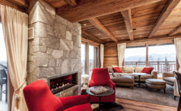 Cozy living room with red chairs, a stone fireplace, and large windows offering mountain views, featuring wooden beams and soft curtains for a warm ambiance.