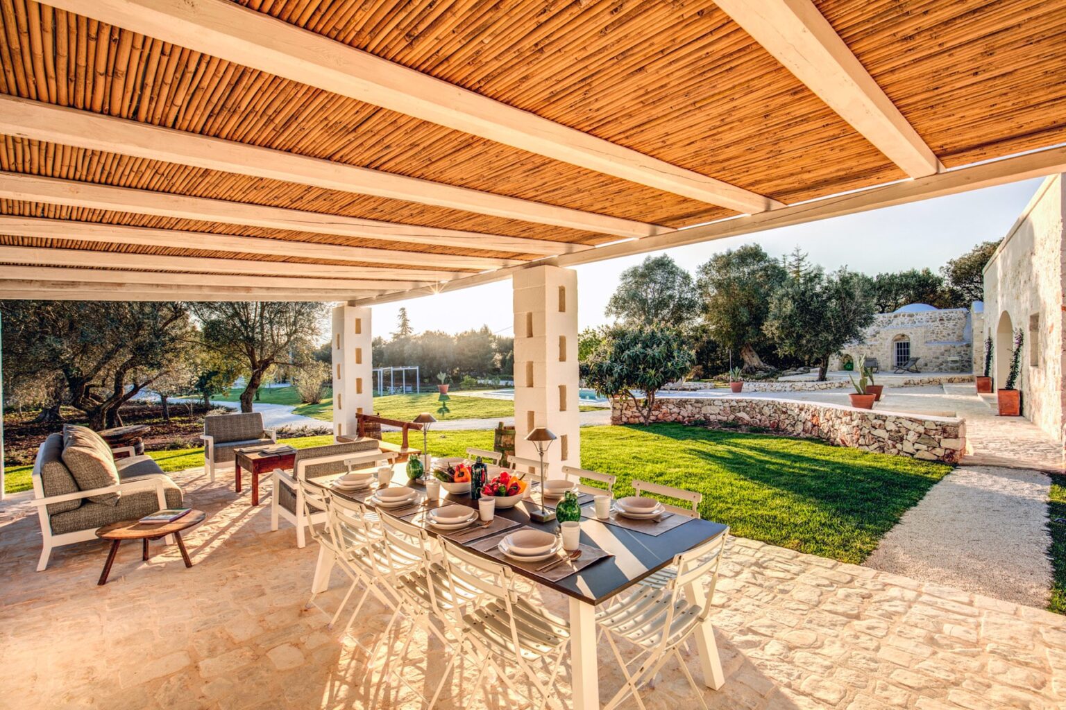Elegant outdoor dining area with a wooden pergola, featuring a stylish table set for a meal, surrounded by comfortable seating and lush green landscaping. Ideal for relaxing gatherings in a serene setting. | Only on SopranoVillas