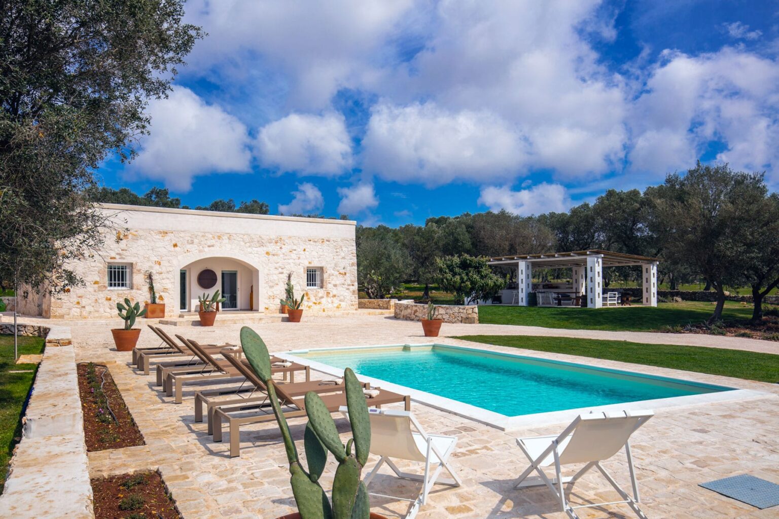 Beautiful outdoor pool area with loungers beside a charming stone villa, surrounded by lush greenery and olive trees under a bright blue sky. Ideal for relaxation and outdoor gatherings. | Only on SopranoVillas