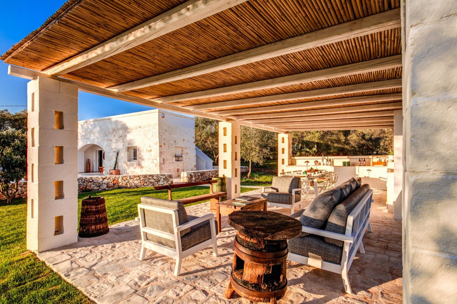 Outdoor seating area with comfortable furniture under a wooden pergola, featuring a rustic stone floor and surrounded by lush greenery, ideal for relaxation and entertaining. | Only on SopranoVillas