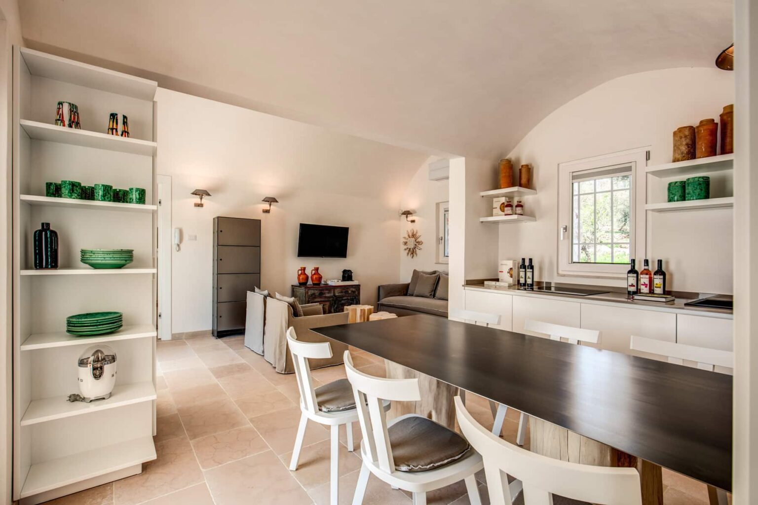 Modern kitchen interior featuring a sleek dining table, white chairs, and open shelves with green dishware. Bright and airy space with a comfortable seating area, television, and natural light from windows. Ideal for entertaining and family gatherings. | Only on SopranoVillas