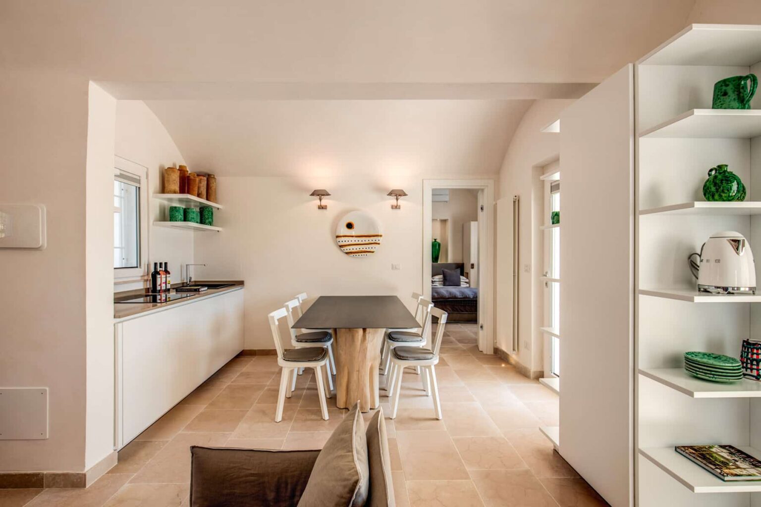 Modern kitchen and dining area with a sleek design, featuring a long table, white cabinetry, and green decorative accents. Natural light fills the space, highlighting the minimalist decor and inviting atmosphere. | Only on SopranoVillas
