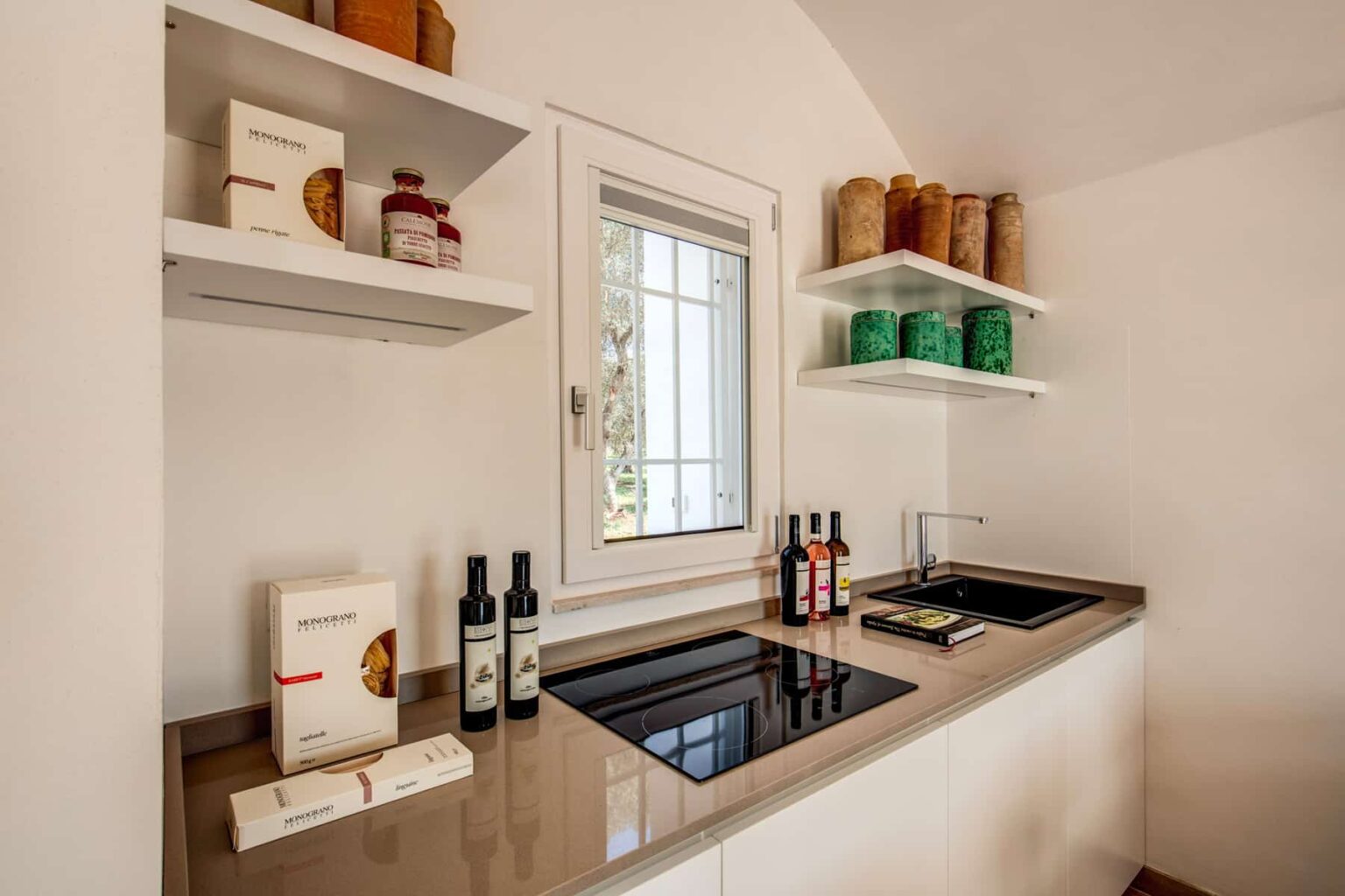 Modern kitchen interior featuring a sleek countertop with a built-in stove, organized shelves displaying olive oil, pasta, and spices, and a window allowing natural light to brighten the space. | Only on SopranoVillas