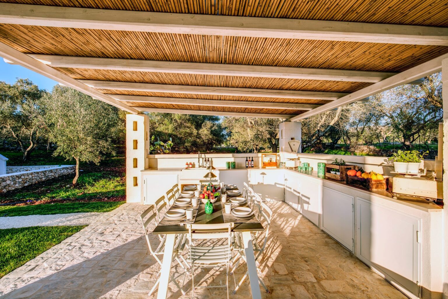 Outdoor kitchen and dining area with modern furnishings, surrounded by greenery and olive trees, featuring a large table set for a meal, under a wooden pergola. | Only on SopranoVillas