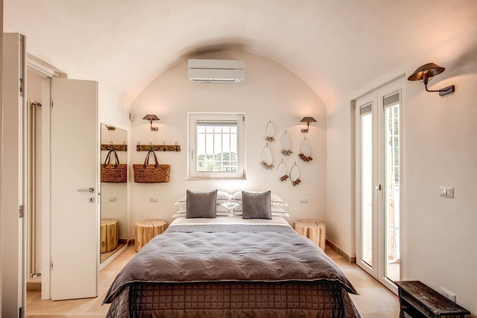 Cozy modern bedroom featuring a double bed with gray bedding, minimalist decor, and natural light from large windows. Includes stylish wall sconces, wooden storage solutions, and an air conditioning unit for comfort. | Only on SopranoVillas