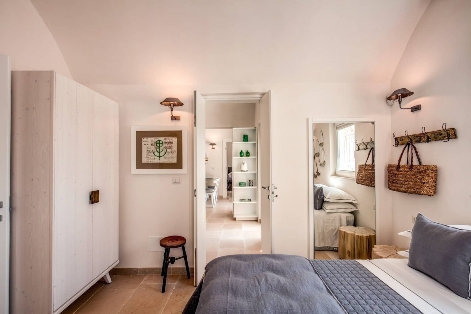 Cozy bedroom with a neutral color palette, featuring a comfortable bed, modern wall sconces, and a small stool. The room has a door leading to a stylish bathroom, with additional decorative elements like a wall art piece and woven baskets, creating a warm and inviting atmosphere. | Only on SopranoVillas