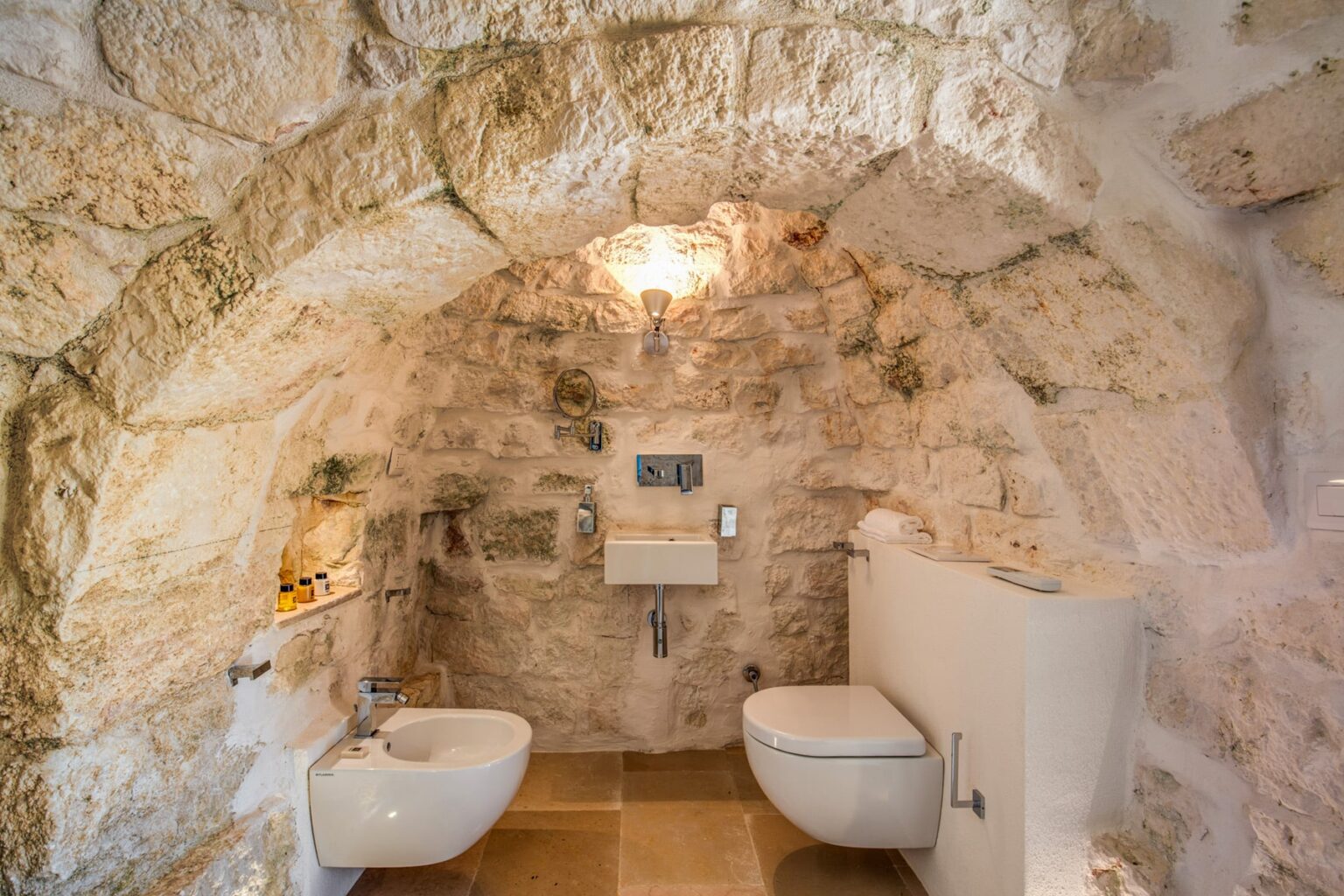 Modern bathroom design featuring a compact toilet and bidet in a rustic stone archway setting, illuminated by a stylish chandelier. | Only on SopranoVillas