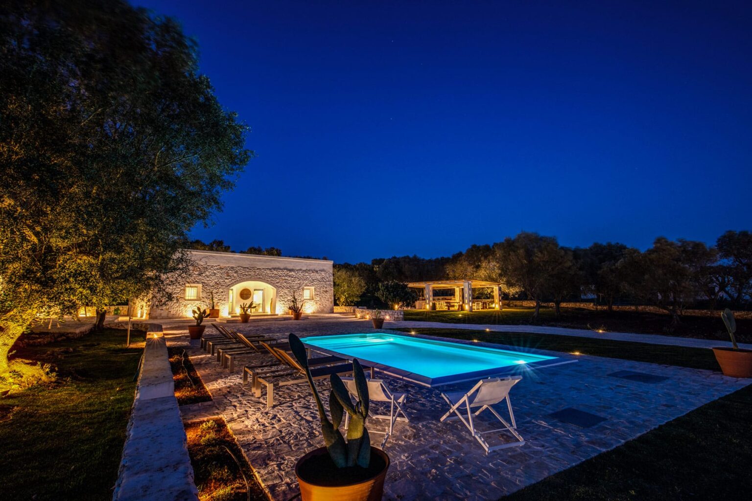 Luxurious outdoor pool area illuminated at night, featuring a serene garden, lounge chairs, and a beautifully designed stone building in the background, perfect for relaxation and entertainment. | Only on SopranoVillas
