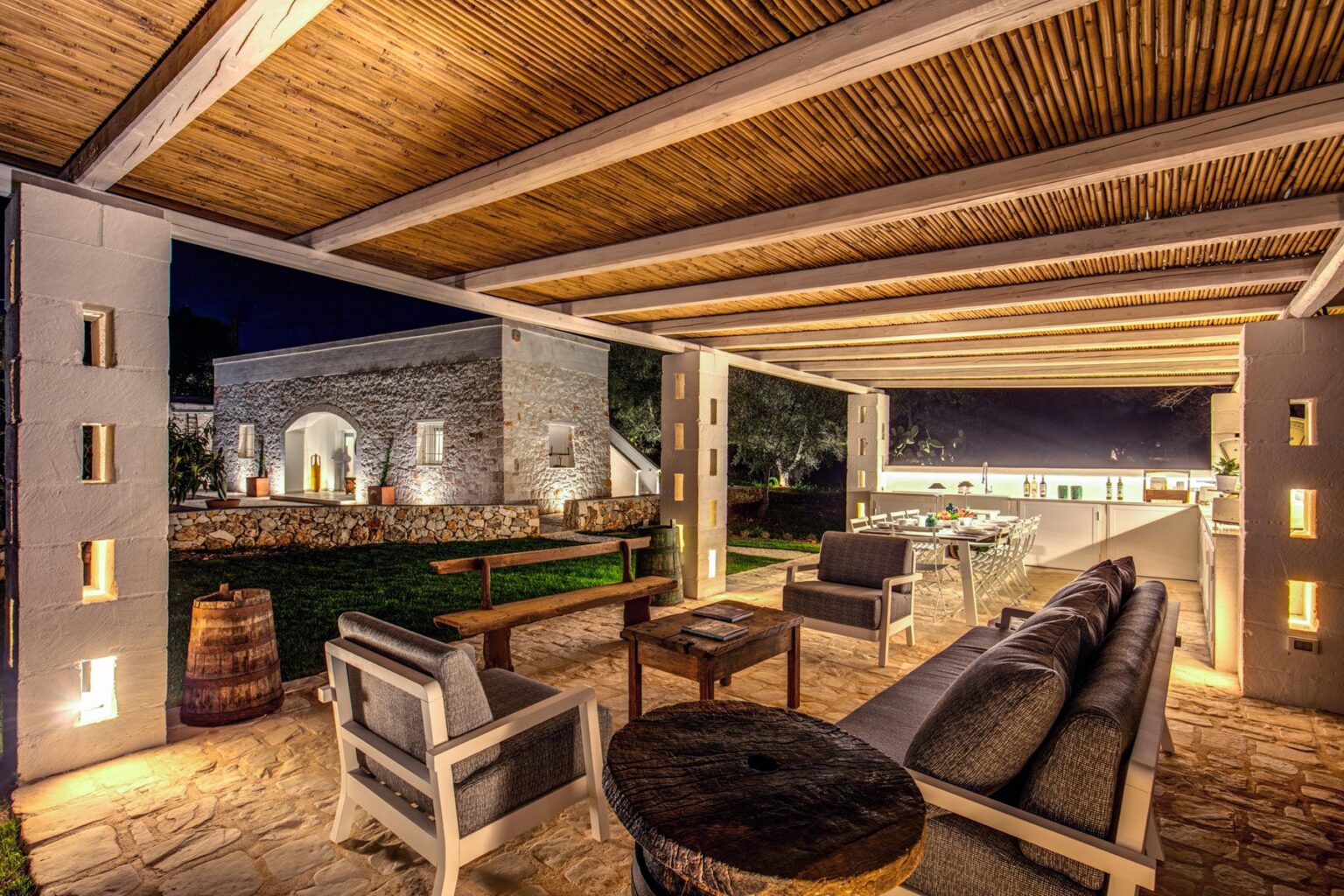 Modern outdoor living space featuring comfortable seating and ambient lighting, showcasing a stylish patio design at night with a view of landscaped gardens. | Only on SopranoVillas
