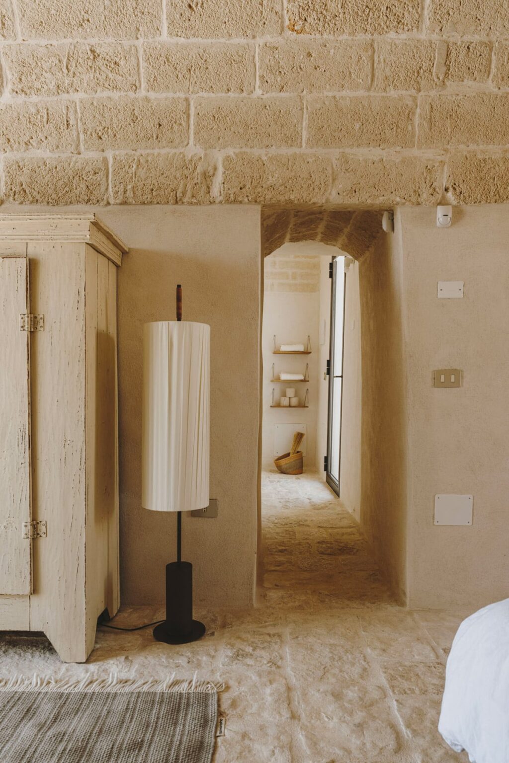 Interior view of a softly lit hallway featuring a stylish floor lamp, textured walls, and an archway leading to a serene space with minimal decor, showcasing a blend of modern and rustic design elements. | Only on SopranoVillas