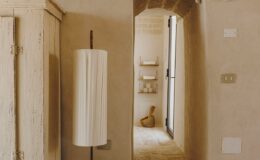 Interior view of a softly lit hallway featuring a stylish floor lamp, textured walls, and an archway leading to a serene space with minimal decor, showcasing a blend of modern and rustic design elements.