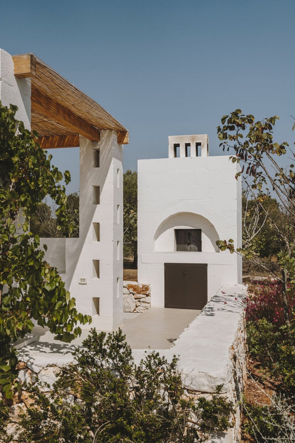 Modern white architecture featuring a minimalist design with distinct geometric shapes, surrounded by greenery. The structure includes a prominent chimney and an arched doorway, reflecting contemporary style in a serene outdoor setting. | Only on SopranoVillas