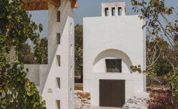 Modern white architecture featuring a minimalist design with distinct geometric shapes, surrounded by greenery. The structure includes a prominent chimney and an arched doorway, reflecting contemporary style in a serene outdoor setting.