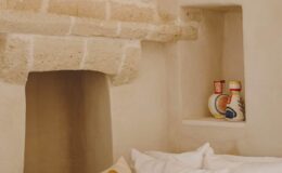 Cozy interior of a rustic room featuring a stone fireplace, soft bedding, and decorative pottery displayed in a niche, highlighting a warm and inviting atmosphere.