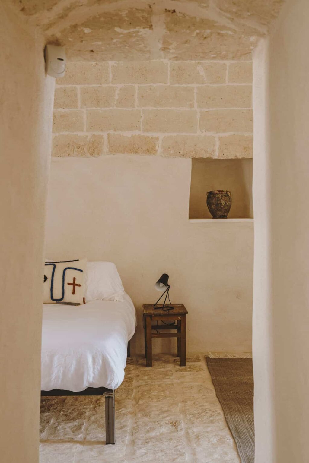 Cozy bedroom interior featuring a white bed with decorative pillows, a wooden nightstand with a lamp, and a decorative vase on a shelf, showcasing a minimalist design with warm, neutral tones. | Only on SopranoVillas