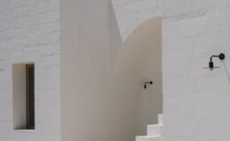 Modern white architecture featuring a textured wall, arched entrance, and minimalist design elements, illuminated by stylish outdoor sconces.