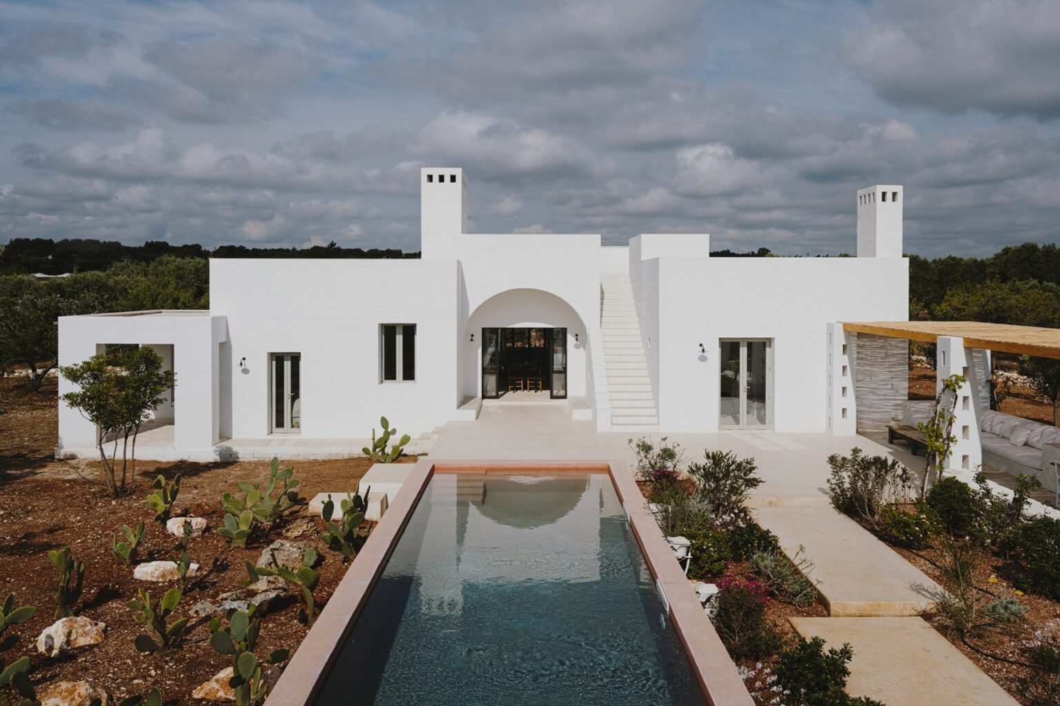Modern white house with an arched entrance, surrounded by landscaped gardens and a reflective pool, under a cloudy sky. | Only on SopranoVillas