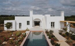 Modern white house with an arched entrance, surrounded by landscaped gardens and a reflective pool, under a cloudy sky.