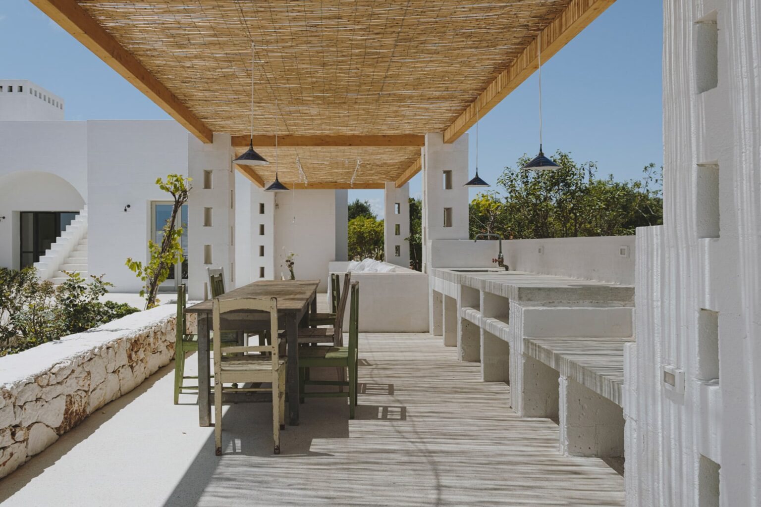Outdoor dining area featuring a wooden table and benches under a thatched roof, surrounded by white walls and greenery, ideal for alfresco meals and relaxation. | Only on SopranoVillas