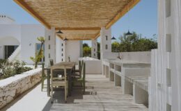 Outdoor dining area featuring a wooden table and benches under a thatched roof, surrounded by white walls and greenery, ideal for alfresco meals and relaxation.
