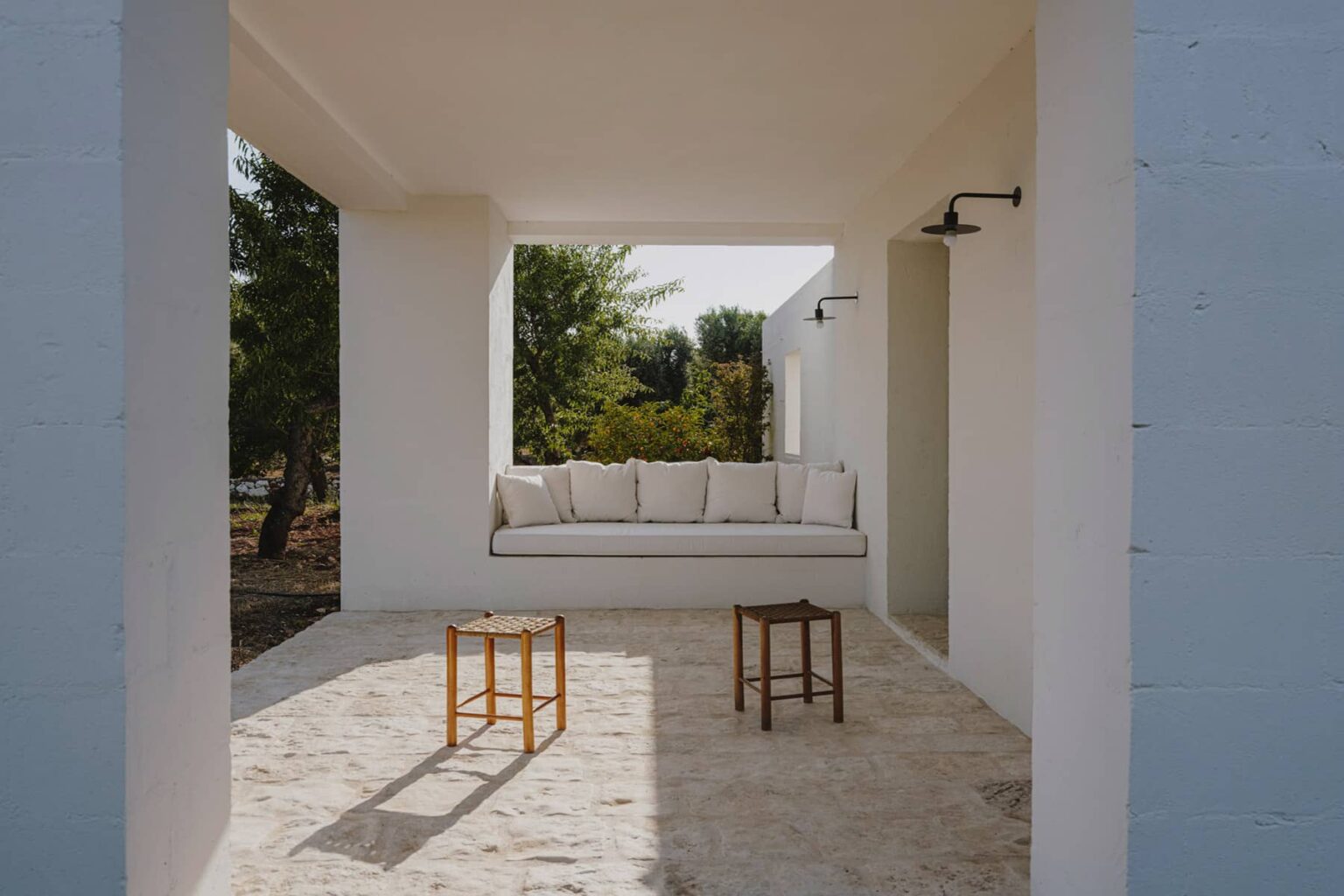 Modern outdoor living space featuring a white minimalist structure, a comfortable built-in bench with cushions, and two wooden stools on a stone floor, surrounded by greenery. | Only on SopranoVillas
