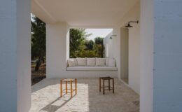 Modern outdoor living space featuring a white minimalist structure, a comfortable built-in bench with cushions, and two wooden stools on a stone floor, surrounded by greenery.