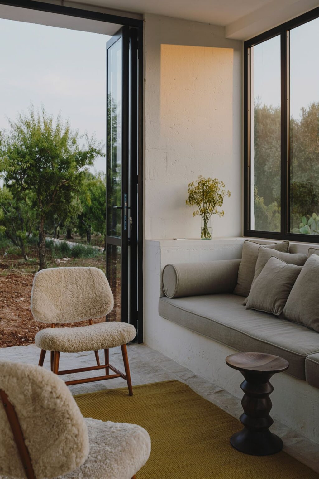 Cozy interior space featuring a comfortable gray sofa with decorative pillows, a soft-textured chair, and a vase of flowers on a shelf, surrounded by large windows that offer views of a lush outdoor landscape. | Only on SopranoVillas