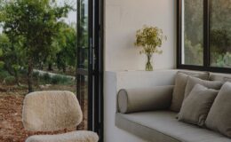 Cozy interior space featuring a comfortable gray sofa with decorative pillows, a soft-textured chair, and a vase of flowers on a shelf, surrounded by large windows that offer views of a lush outdoor landscape.