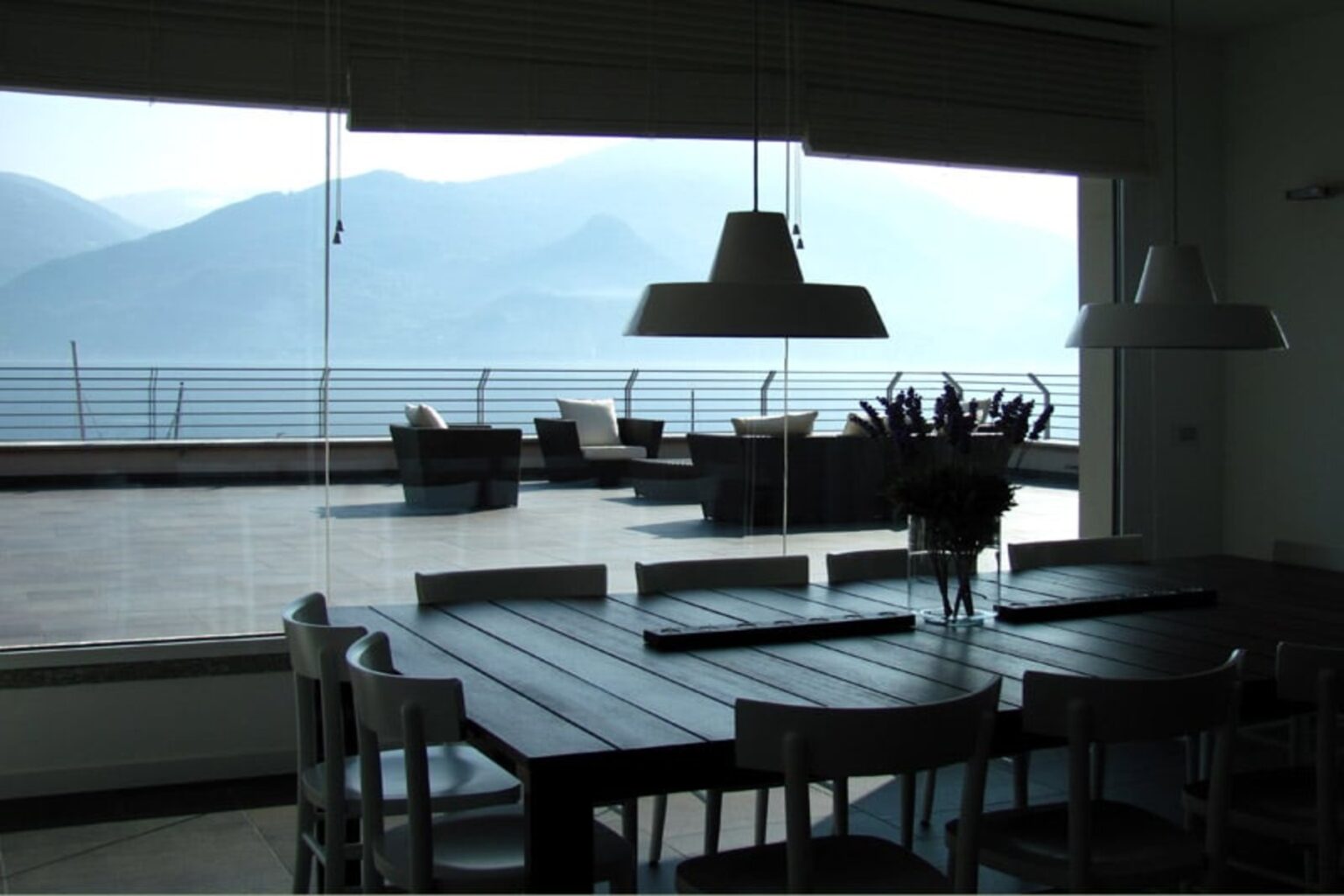Modern dining area with a wooden table and chairs, featuring large windows that offer a view of mountains and a terrace. Natural light floods the space, creating a serene atmosphere. | Only on SopranoVillas