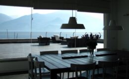 Modern dining area with a wooden table and chairs, featuring large windows that offer a view of mountains and a terrace. Natural light floods the space, creating a serene atmosphere.