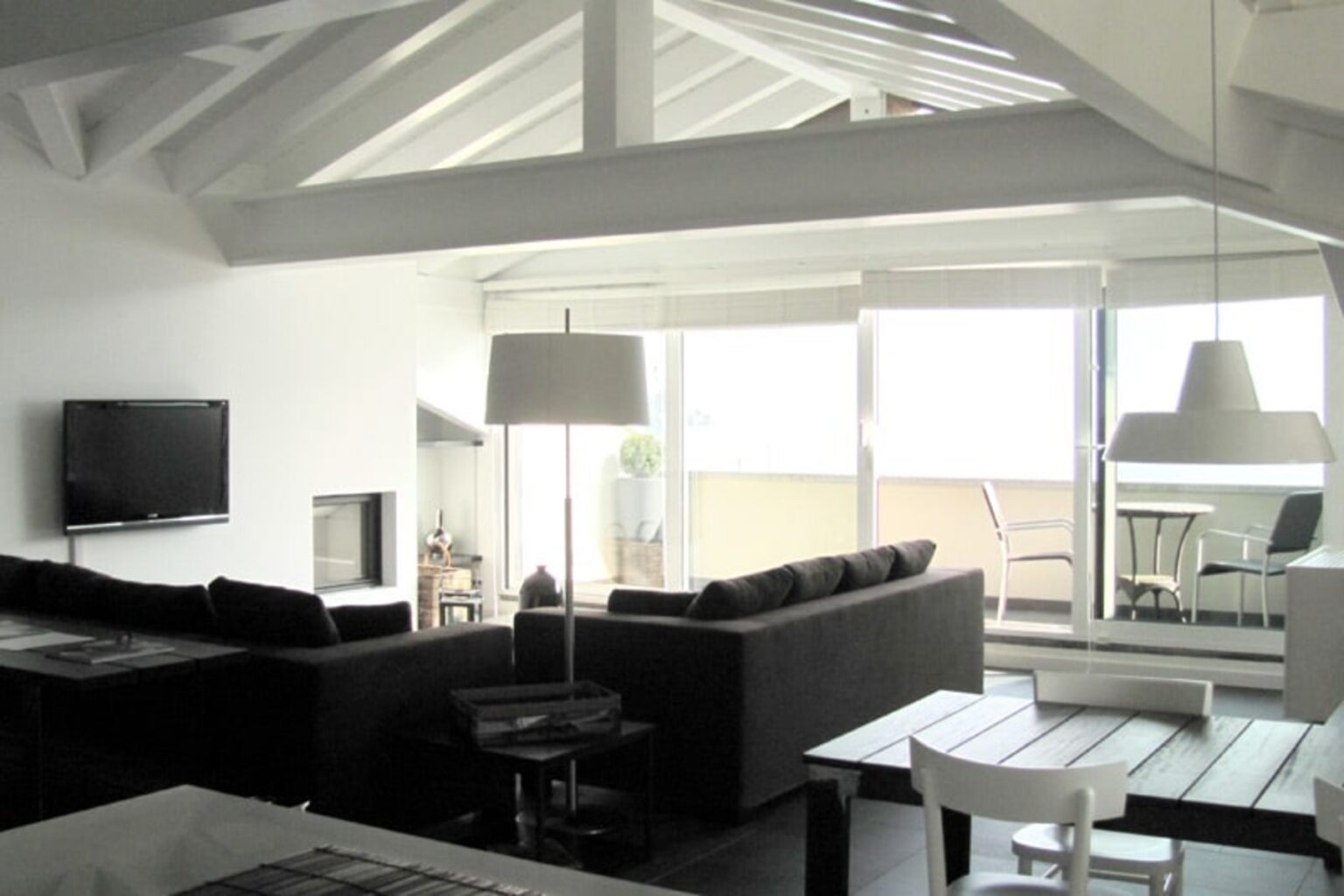 Modern living room with high ceilings, featuring a black sectional sofa, a television, and large windows that provide natural light. The space includes a dining area with a table and chairs, and a minimalist design with white and neutral tones. | Only on SopranoVillas
