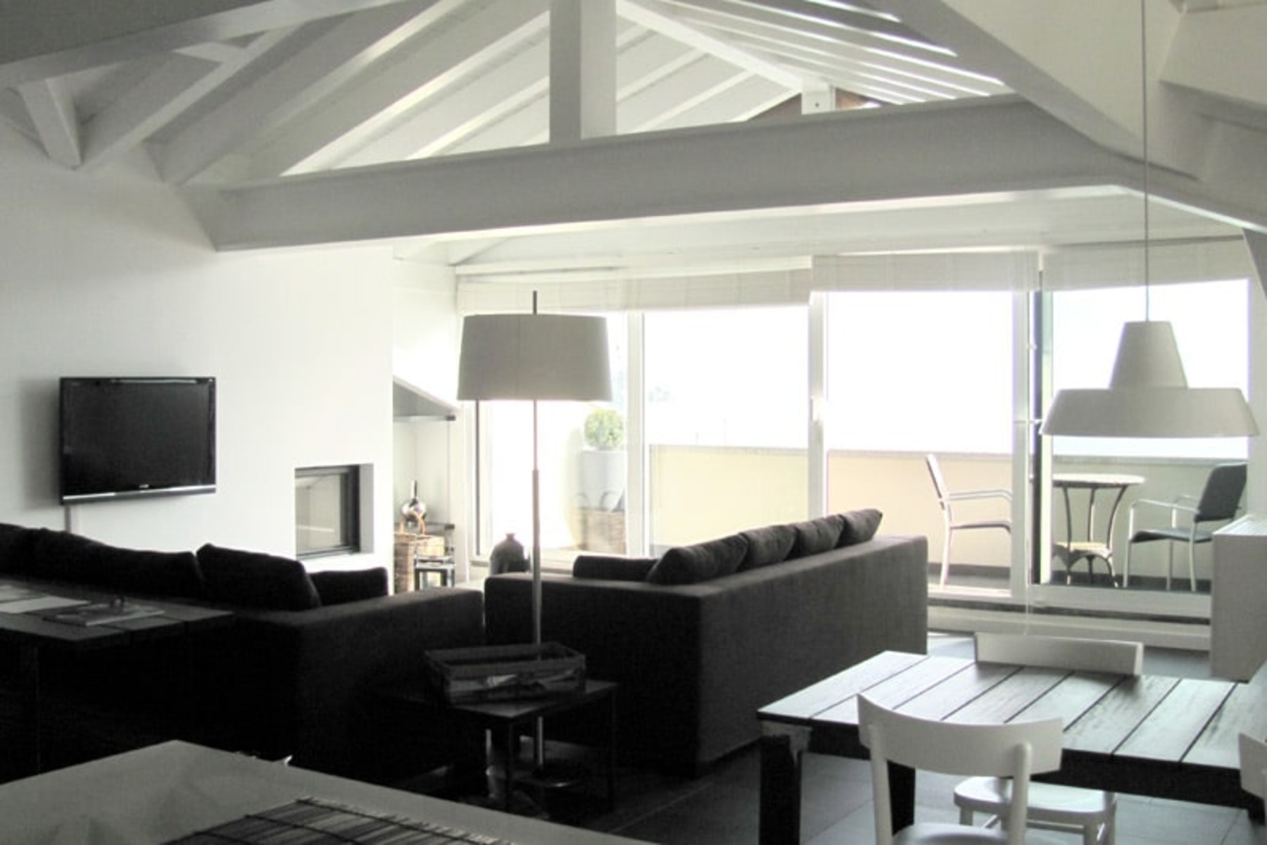 Modern living room with high ceilings, featuring a black sectional sofa, a television, and large windows that provide natural light. The space includes a dining area with a table and chairs, and a minimalist design with white and neutral tones. | Only on SopranoVillas