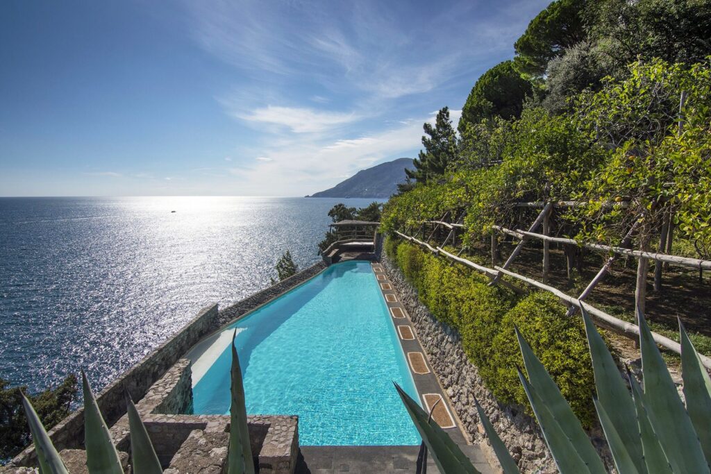 Stunning infinity pool overlooking the serene ocean, surrounded by lush greenery and rocky cliffs, under a bright blue sky. Perfect for luxury vacation relaxation.