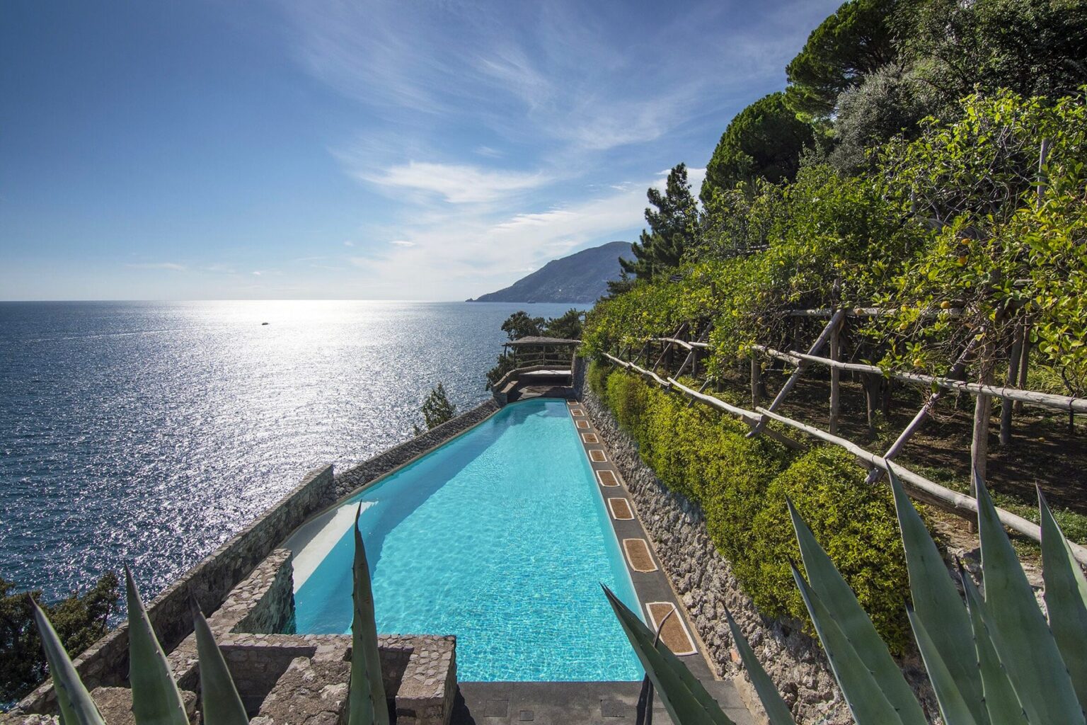 Stunning infinity pool overlooking the serene ocean, surrounded by lush greenery and rocky cliffs, under a bright blue sky. Perfect for luxury vacation relaxation. | Only on SopranoVillas
