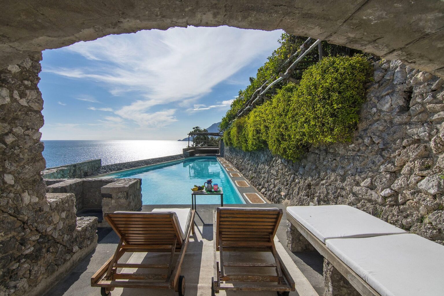 Scenic view of a luxury poolside area with wooden lounge chairs, framed by a stone archway and surrounded by lush greenery, overlooking the shimmering ocean. | Only on SopranoVillas