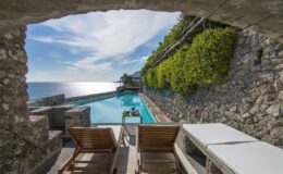Scenic view of a luxury poolside area with wooden lounge chairs, framed by a stone archway and surrounded by lush greenery, overlooking the shimmering ocean.
