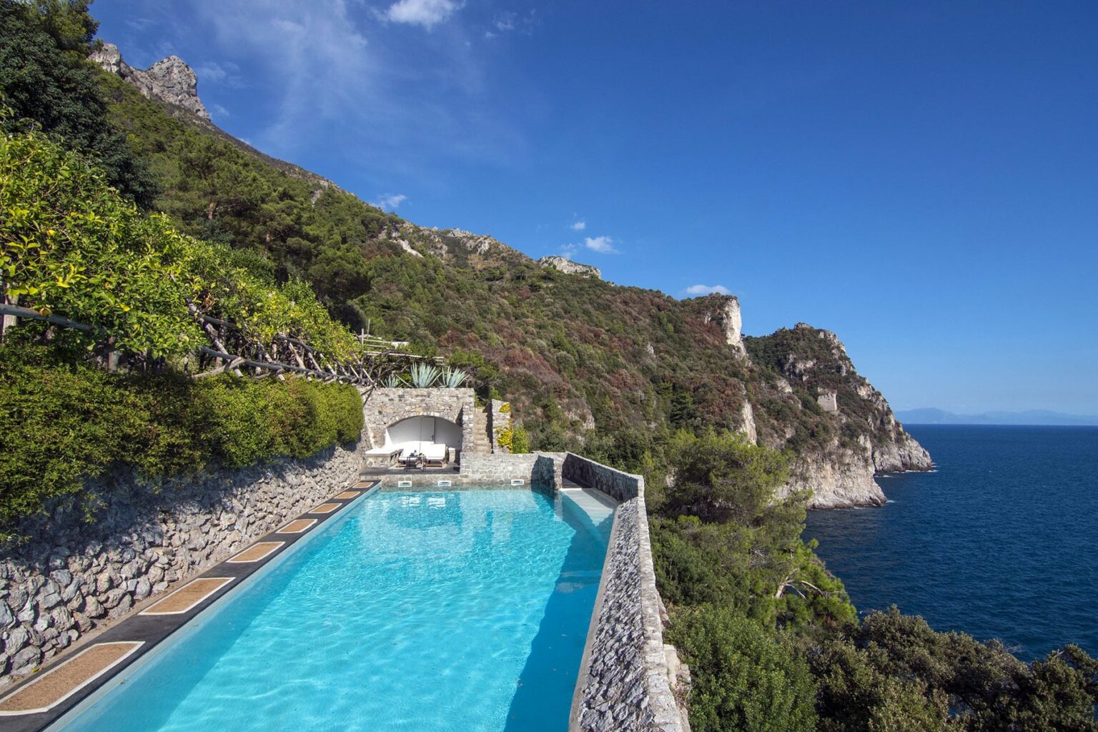 Stunning infinity pool overlooking the ocean, surrounded by lush greenery and rocky cliffs under a clear blue sky. Ideal for luxury vacation destinations or serene getaways. | Only on SopranoVillas