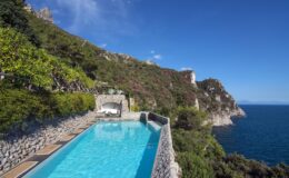 Stunning infinity pool overlooking the ocean, surrounded by lush greenery and rocky cliffs under a clear blue sky. Ideal for luxury vacation destinations or serene getaways.