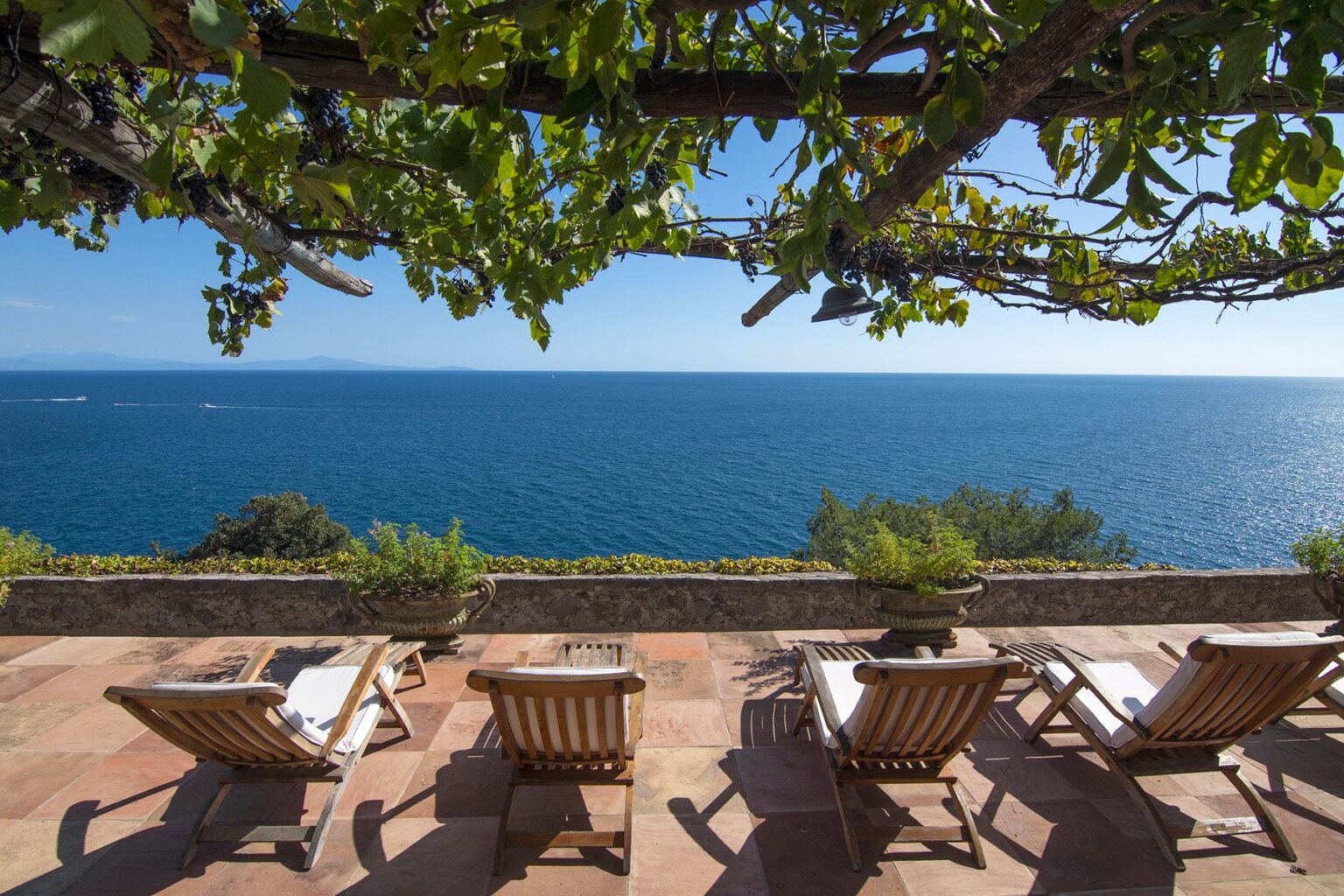 Scenic view of a tranquil ocean from a terrace featuring four wooden lounge chairs under a vine-covered pergola, ideal for relaxation and enjoying coastal sunsets. | Only on SopranoVillas