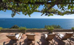 Scenic view of a tranquil ocean from a terrace featuring four wooden lounge chairs under a vine-covered pergola, ideal for relaxation and enjoying coastal sunsets.