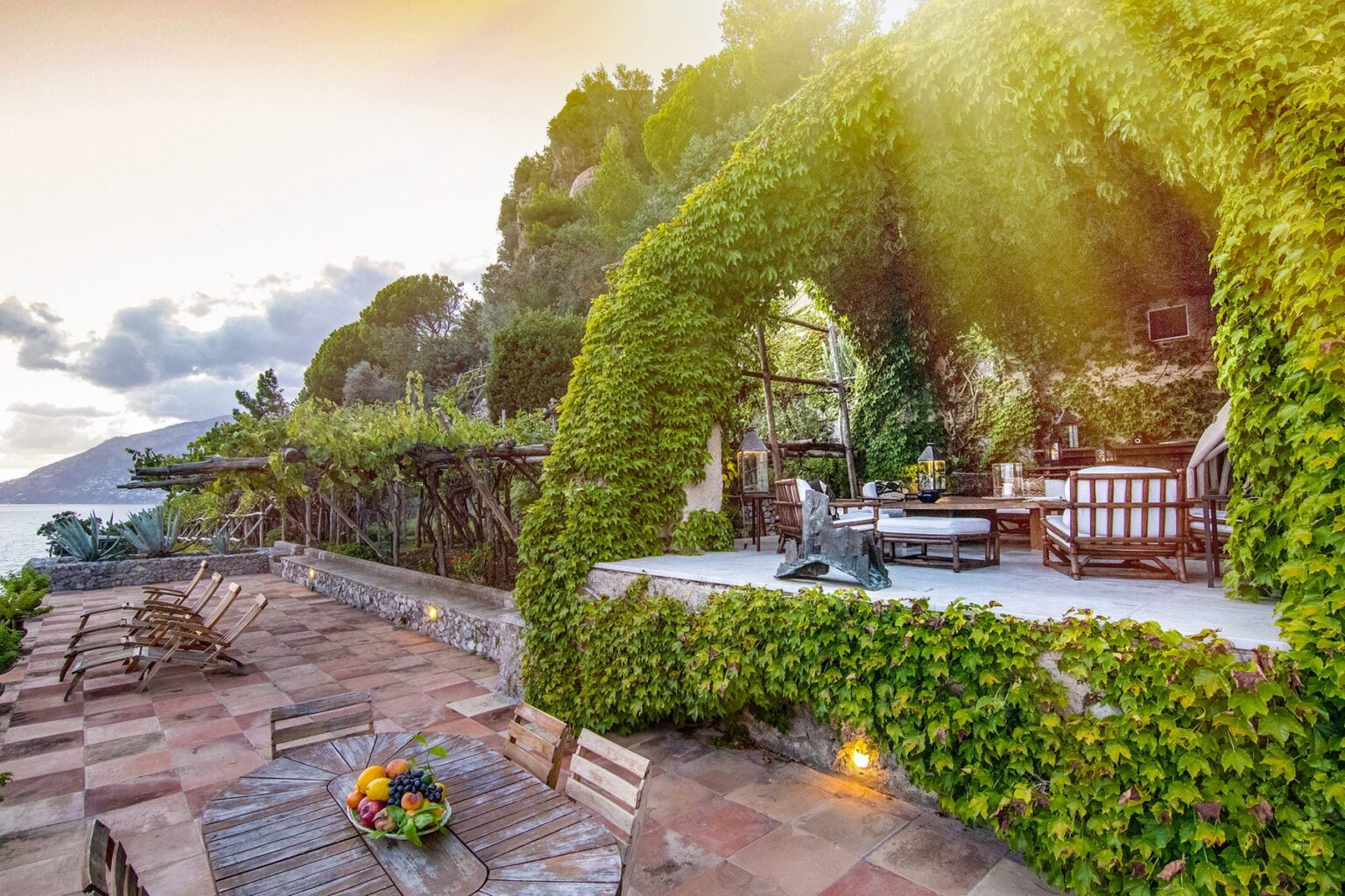 Lush garden patio with a stone pathway, featuring a vine-covered arch and outdoor seating area overlooking a serene landscape at sunset. | Only on SopranoVillas