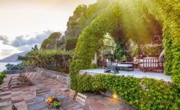 Lush garden patio with a stone pathway, featuring a vine-covered arch and outdoor seating area overlooking a serene landscape at sunset.