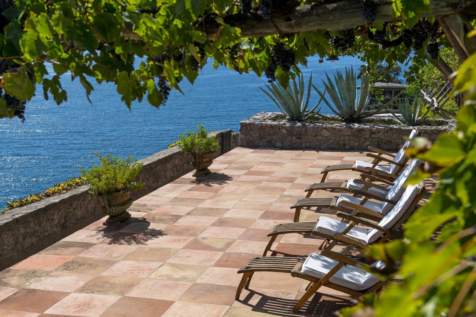 Outdoor patio with wooden lounge chairs overlooking a serene body of water, surrounded by lush greenery and decorative plants. Ideal for relaxation and enjoying scenic views. | Only on SopranoVillas