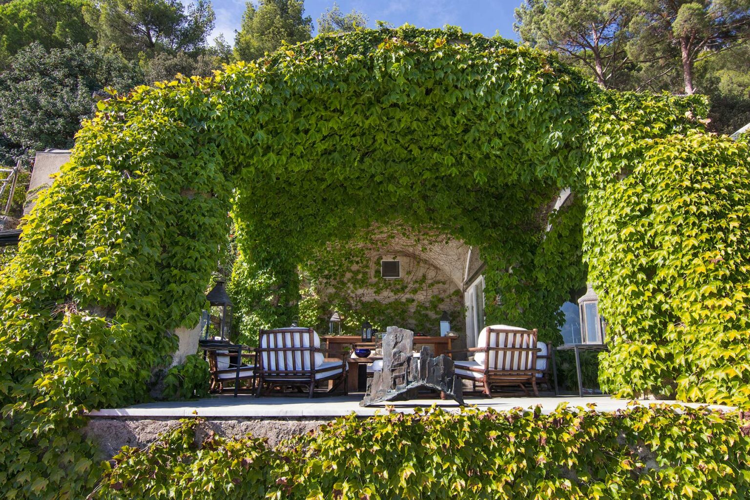 Lush green archway covered in ivy leading to a cozy outdoor seating area with comfortable chairs and a rustic table, surrounded by nature. Ideal for relaxation and enjoying scenic views. | Only on SopranoVillas