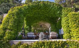 Lush green archway covered in ivy leading to a cozy outdoor seating area with comfortable chairs and a rustic table, surrounded by nature. Ideal for relaxation and enjoying scenic views.