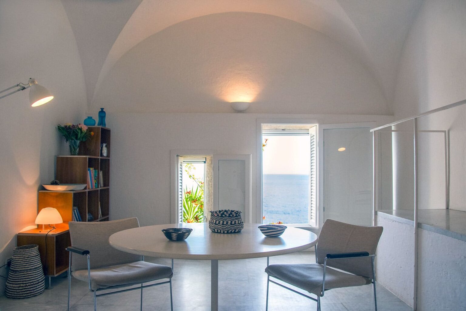 Bright and airy dining area with a round table, modern chairs, and a minimalist design, featuring large windows that let in natural light and a view of greenery. | Only on SopranoVillas