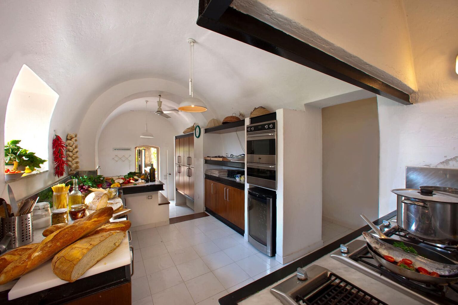 Spacious modern kitchen featuring arched ceilings, wooden cabinetry, stainless steel appliances, and a variety of fresh ingredients on the counter, creating an inviting culinary atmosphere. | Only on SopranoVillas